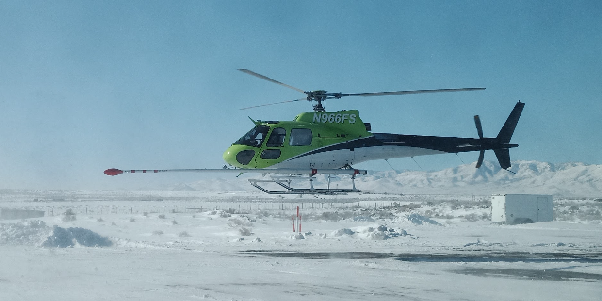 Image shows a helicopter with a red-tipped stinger sticking out front taking off from a snow-covered airport.