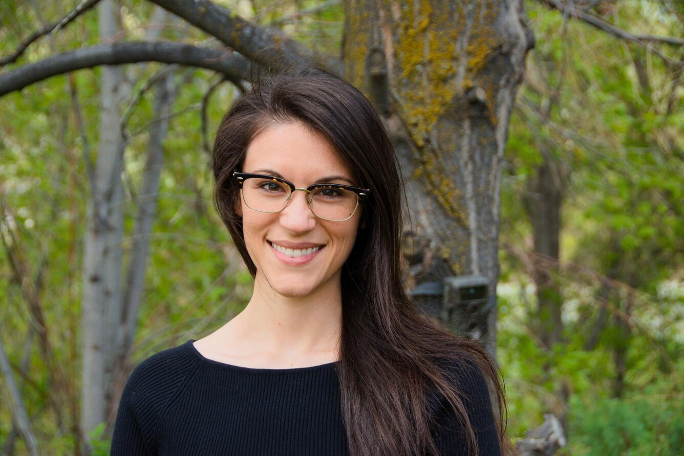 female scientist smiling, with long brown hair, wearing glasses and a black shirt, standing in the woods