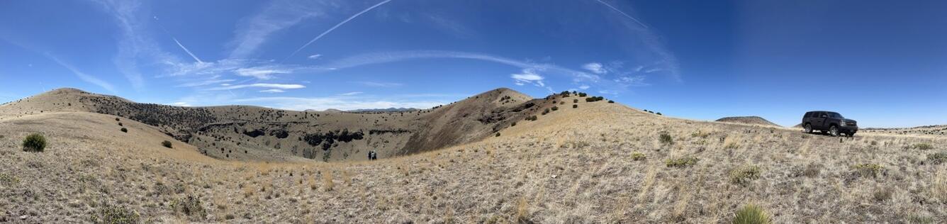 A panoramic photo shows a deep pit with layered crater walls and gentle outer slopes. The walls of the crater are rocky, but the rest of the slopes are covered in gray-tan grass and dark green shrubs. At right, a large SUV is parked on one slope. 
