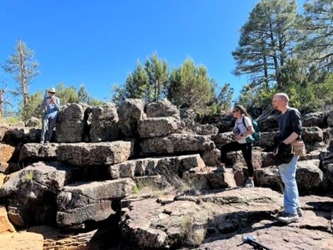 In this photo, three scientists are examining a large pile of slabs of lava. One man wearing a blue button-down shirt and khaki hat is perched on an upper level of the slabs at left, a young woman with braided hair has one foot perched on a slab like a certain pirate at middle right, and a tall bald man in a black shirt stands on top of a pavement of hexagonal lava columns at far right. In the background, scrubby pine trees and bushes grow atop the lava. 
