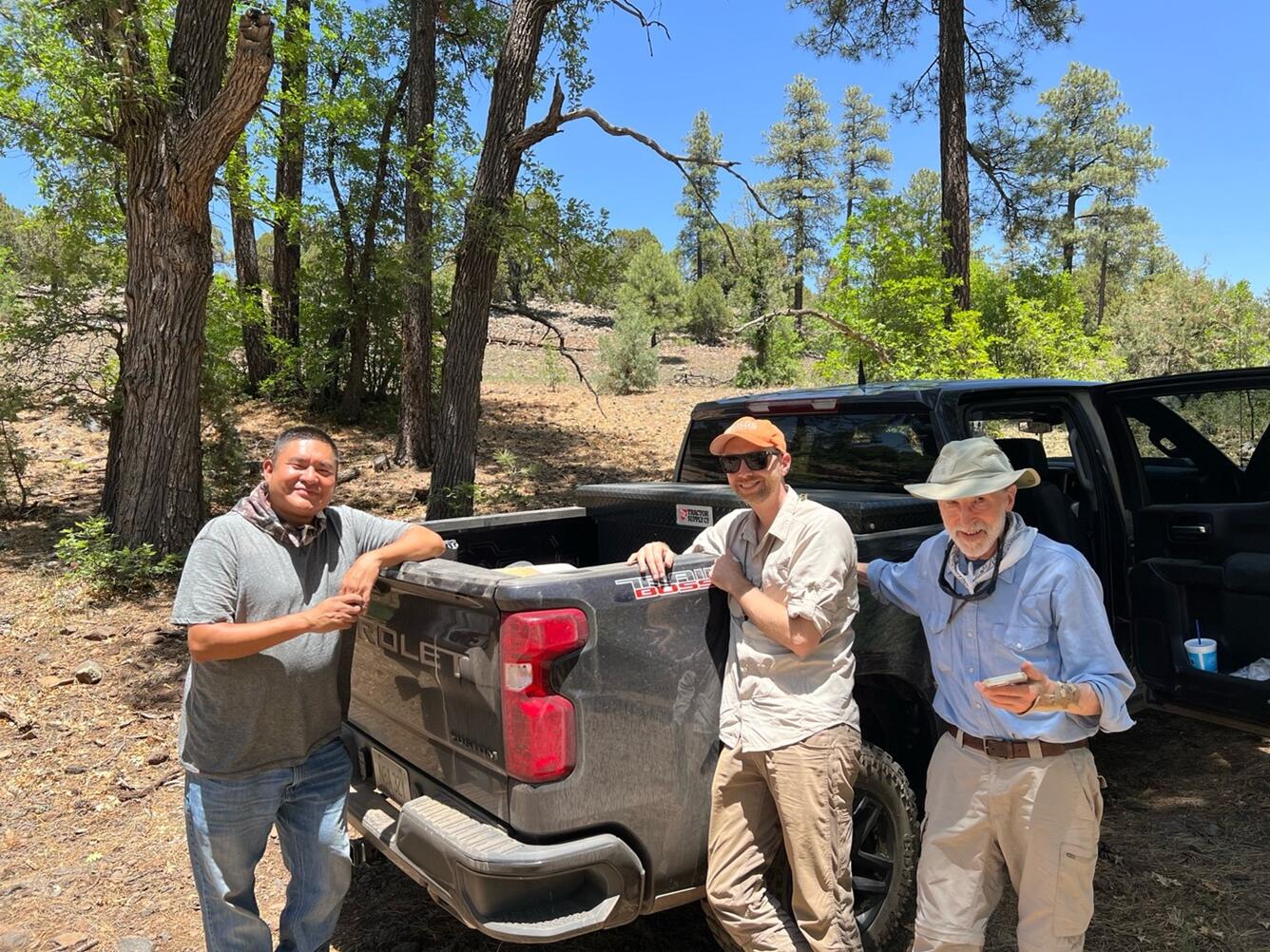 A photograph of three men leaning against the bed of a dusty black truck. The man on the left has tan-colored skin, short dark hair, and is wearing dusty jeans, a gray t-shirt, and a colorful bandanna. The man in the center is wearing khaki pants, a light blue button-down shirt, an orange USGS ballcap, and dark sunglasses. The man on the right is older and wearing khaki pants, a blue button-down shirt, a white bandanna, and a khaki hat, with sunglasses on a lanyard around his neck. 