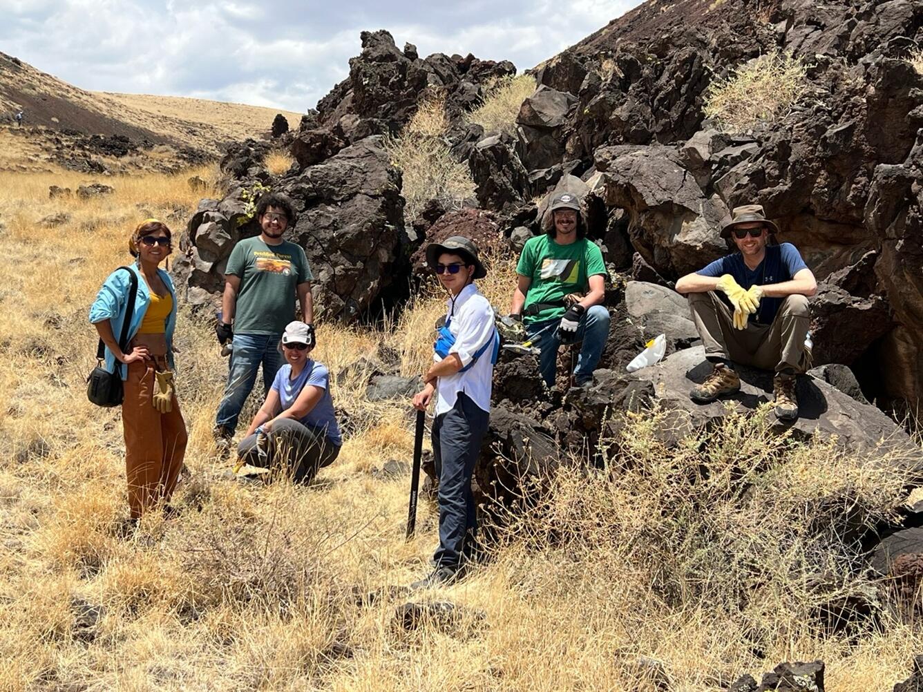 In this photo, a group of six people stand and sit on the edge of a lava flow made up of craggy black boulders. From left to right, a woman wearing brown pants, blue and yellow shirts, and a kerchief; a bearded man wearing jeans and a green t-shirt; a kneeling woman wearing black pants, a blue t-shirt and a khaki ballcap; a man wearing black pants, a white shirt and a gray hat; a man with a moustache wearing jeans, a green t-shirt and a gray ballcap; and a sitting man wearing khaki pants and a blue shirt.