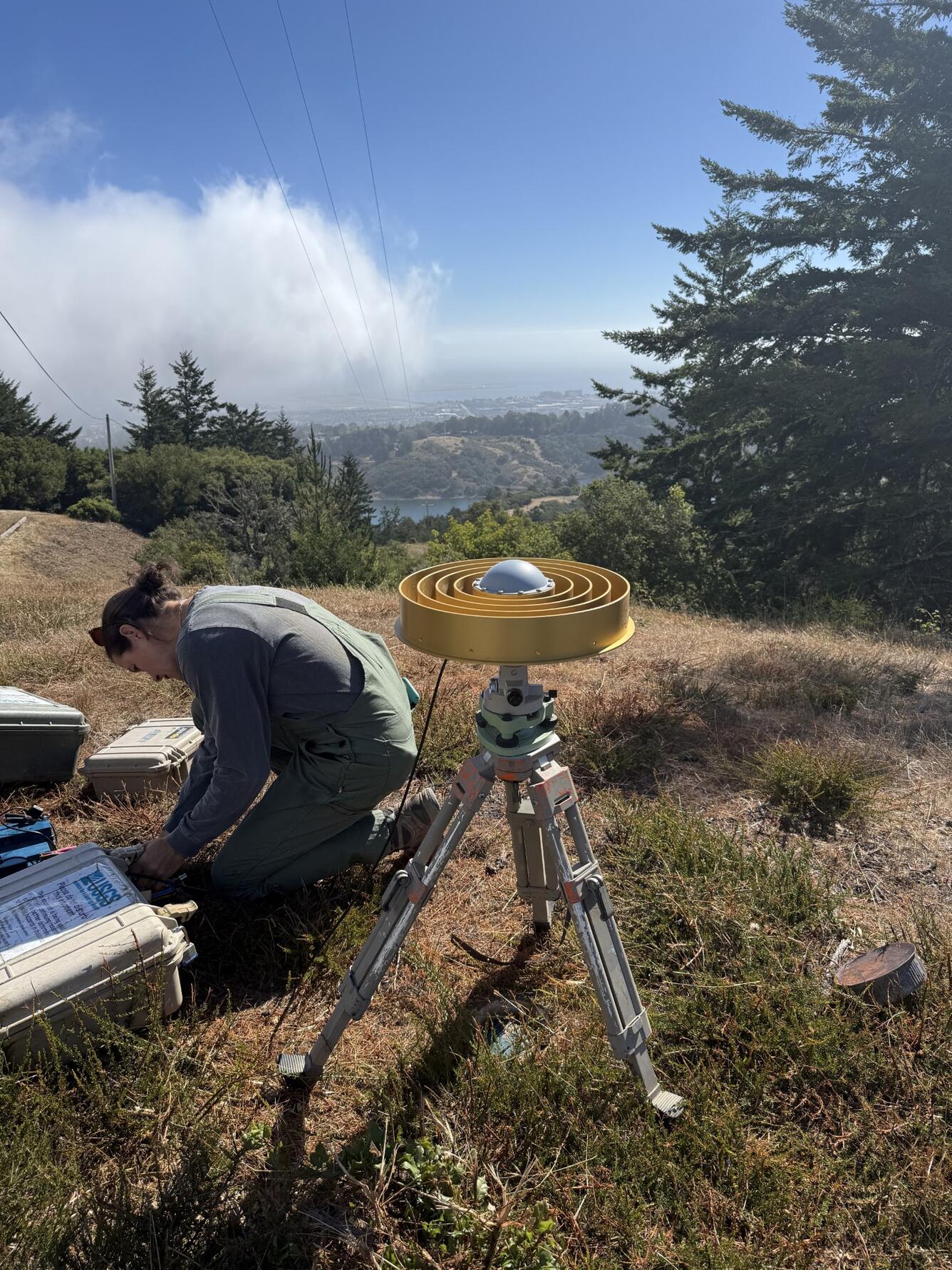 USGS scientist kneels beside gold ring GNSS antenna on a hillside above San Andreas Lake with fog along the distant coast.