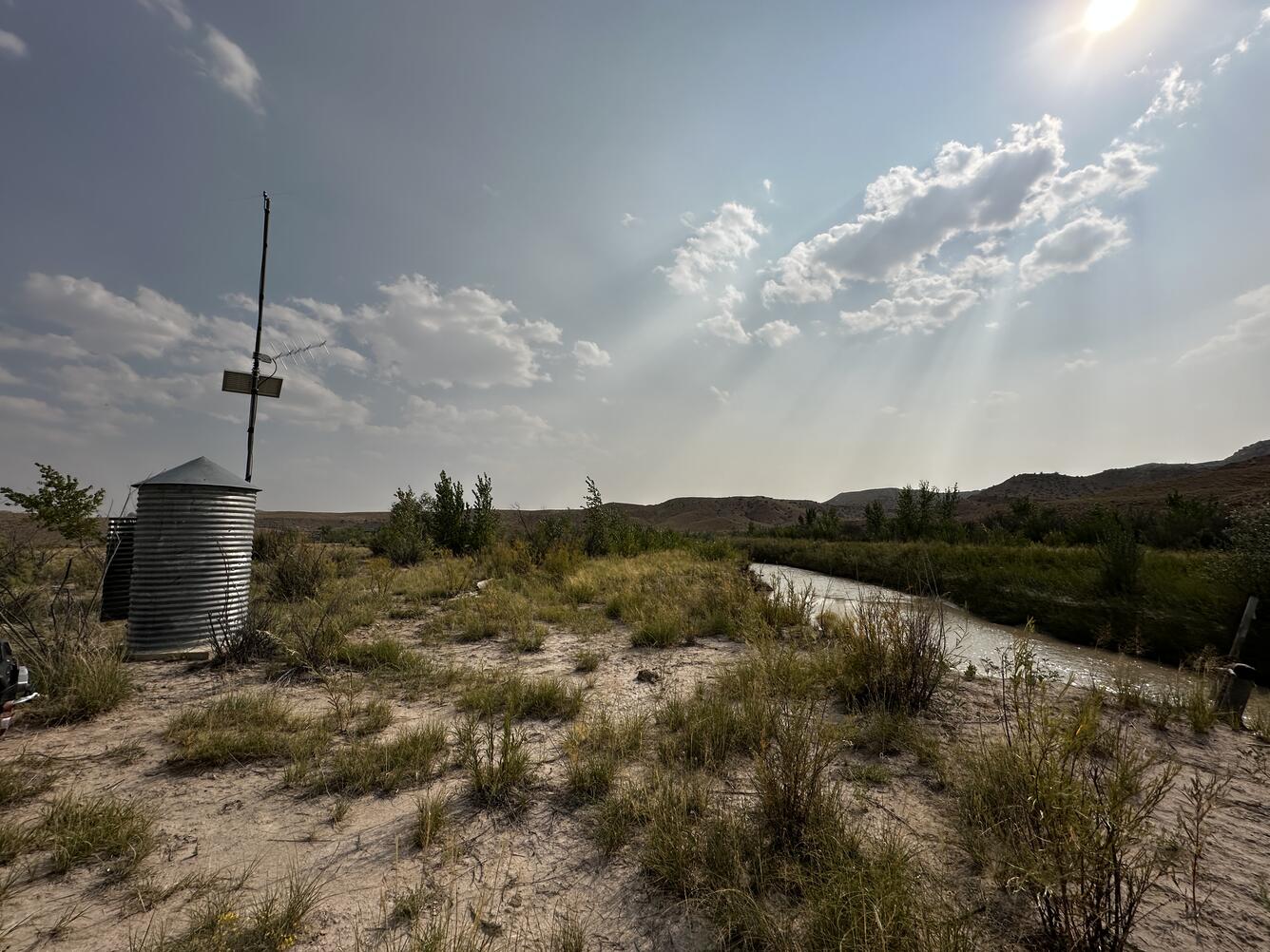 Cylindrical corrugated galvanized steel gage house on flat sparsely vegetated stream bank.