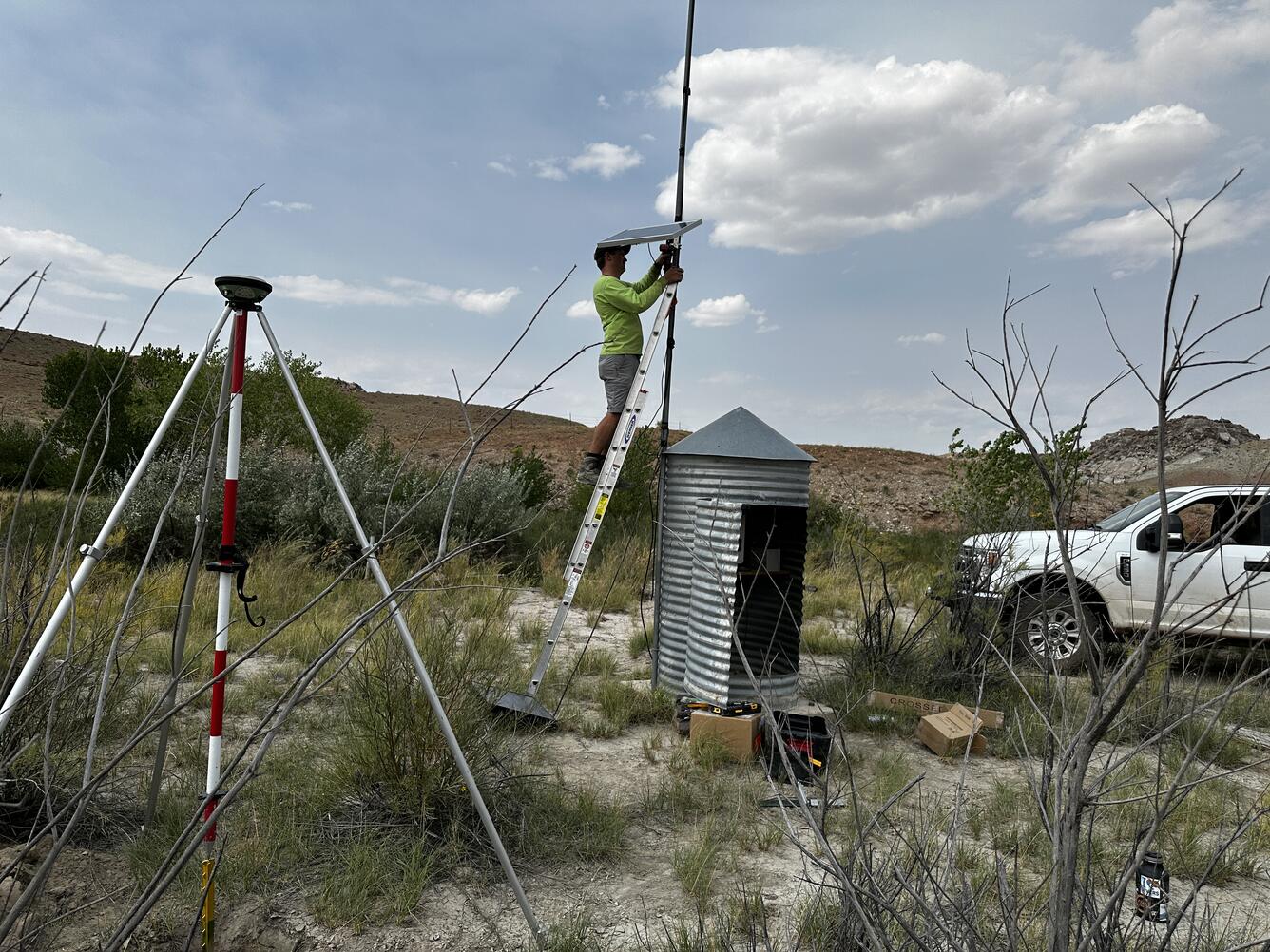 Person on ladder next to cylindrical gage house installing solar panel on pole.