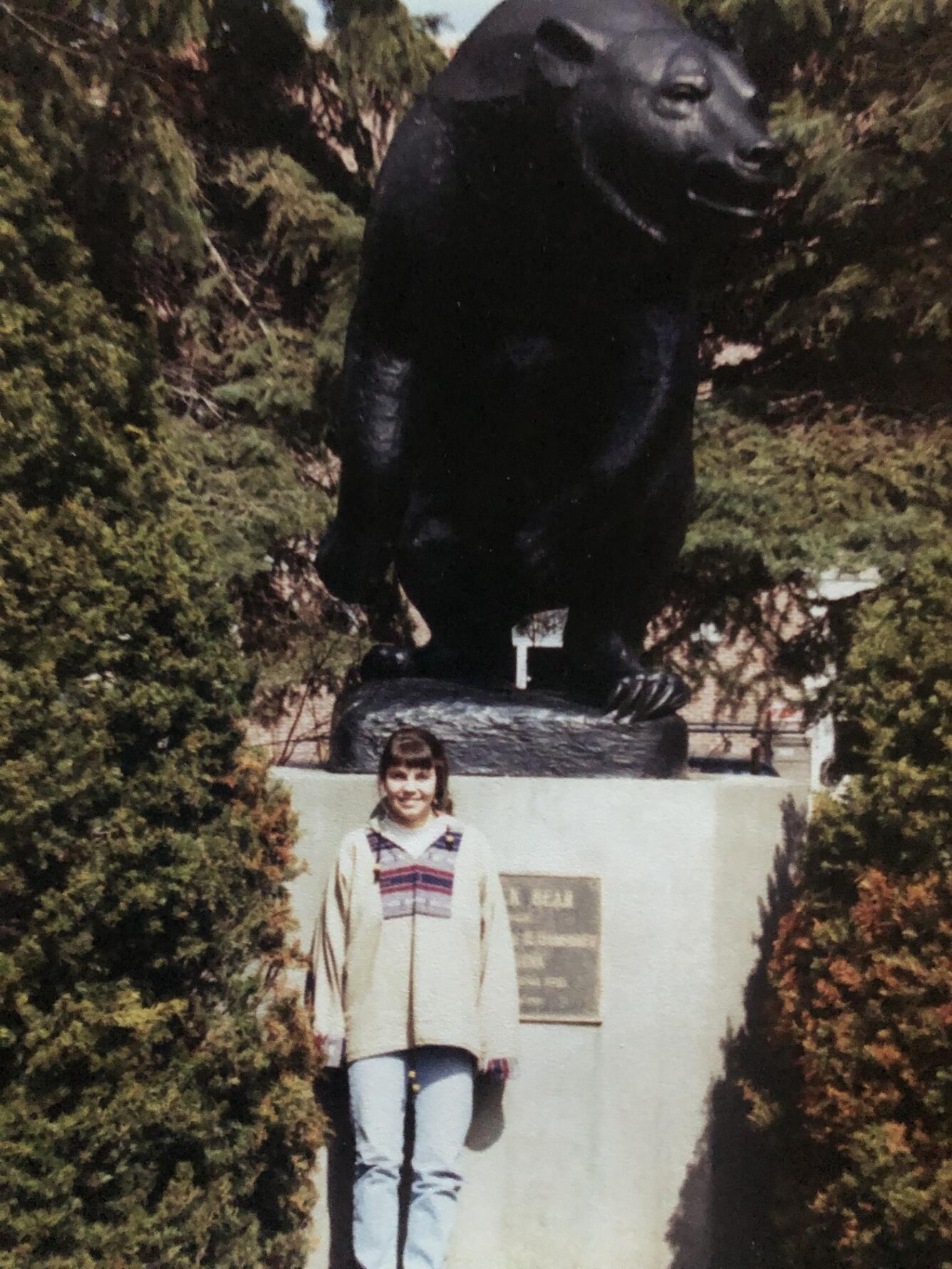 a graduate student in white sweatshirt and blue jeans stands in front of a large bear statue