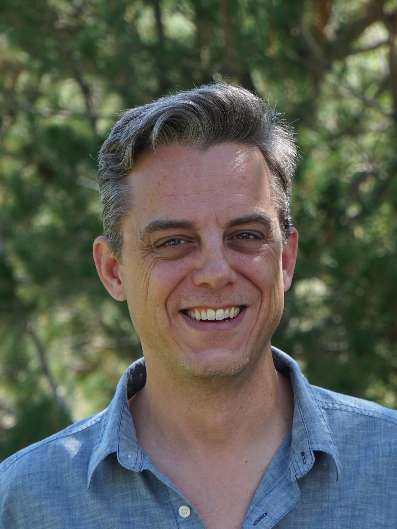 male scientist smiling, with gray black hair, wearing a blue shirt, standing near trees