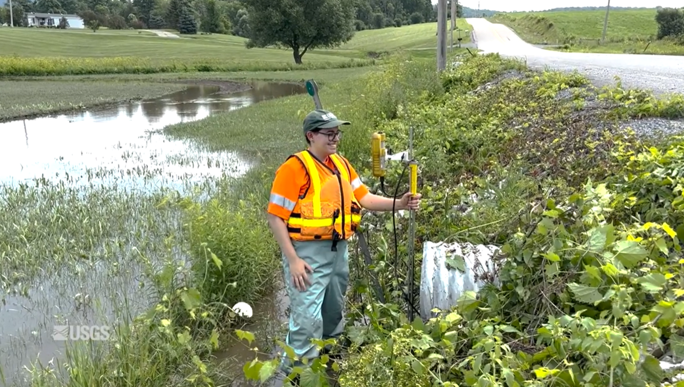 A woman wearing an orange life jacket takes a stream discharge measurement in front of a culvert.