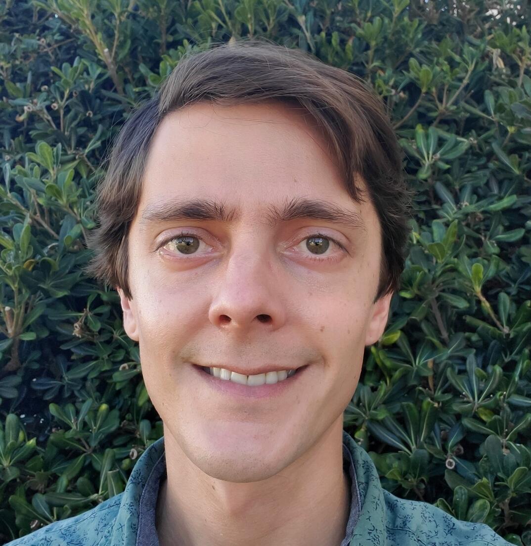 Photograph of a young man with straight brown hair and hazel eyes set against a green leafy backdrop