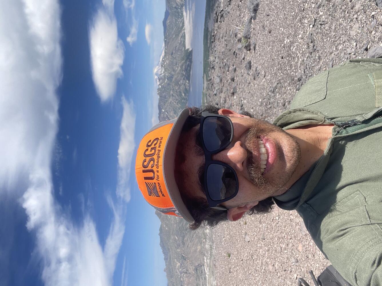 A young man in a green flight suit, orange hat, and dark sunglasses stands on a rocky mountain near a lake. He has brown hair and mustache and fair skin.