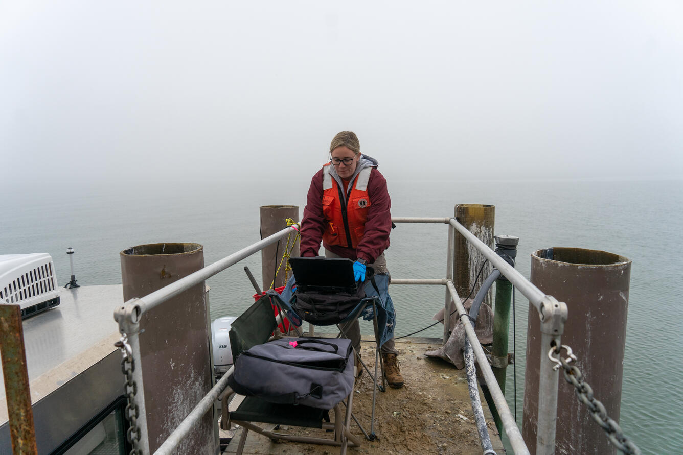 A person rests a computer on a folding chair along a muddy dock.