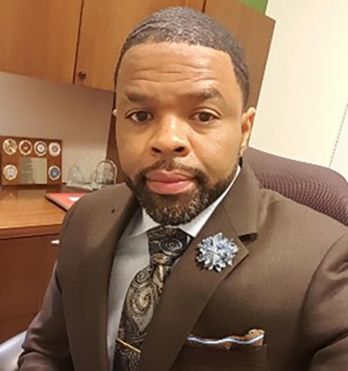 Portrait of Rickey Siggal sitting at a desk in a brown suit and tie smiling with brown cabinets hang on wall behind him.