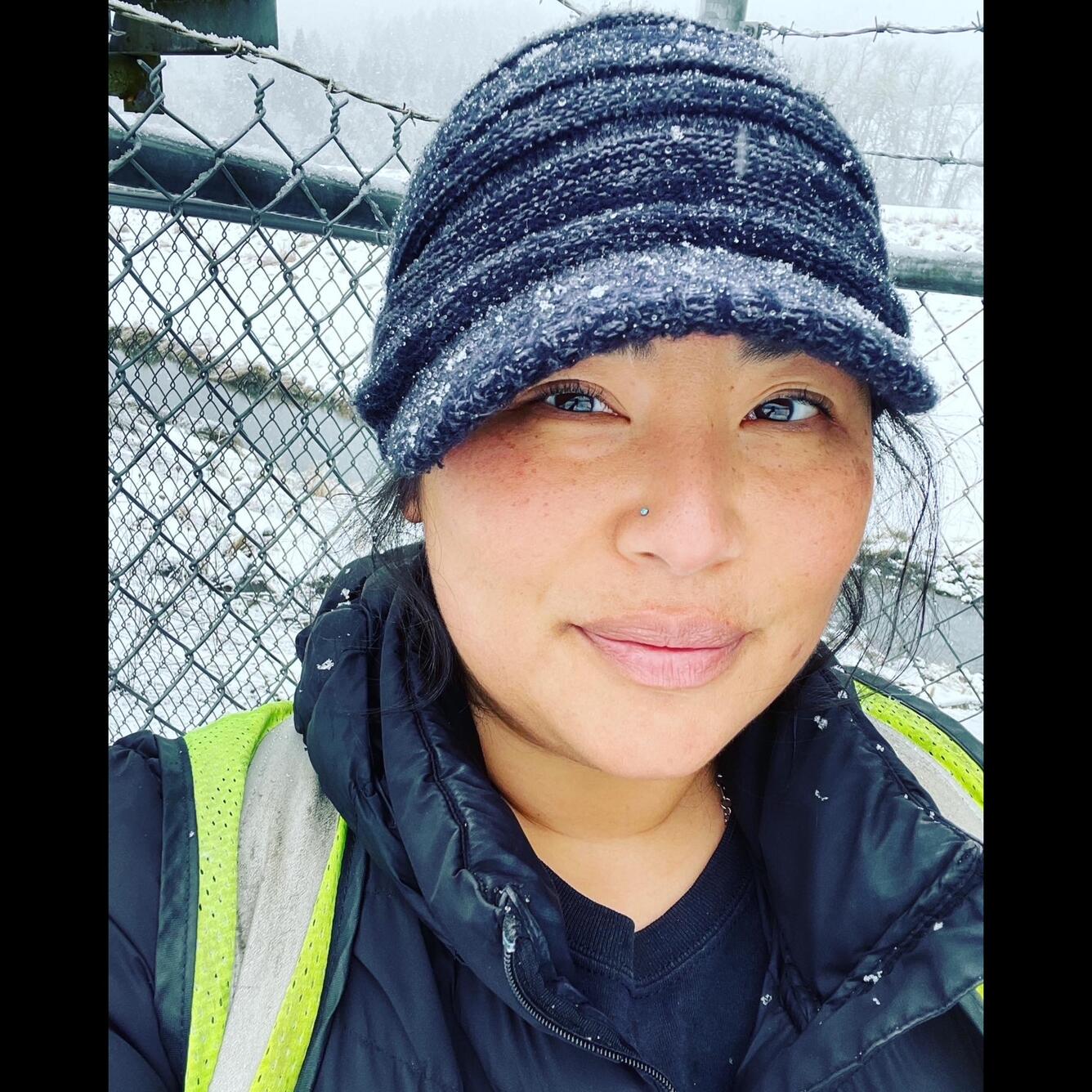 Headshot of a female wearing black snow dusted hat next tp a metal fence on a snowy day