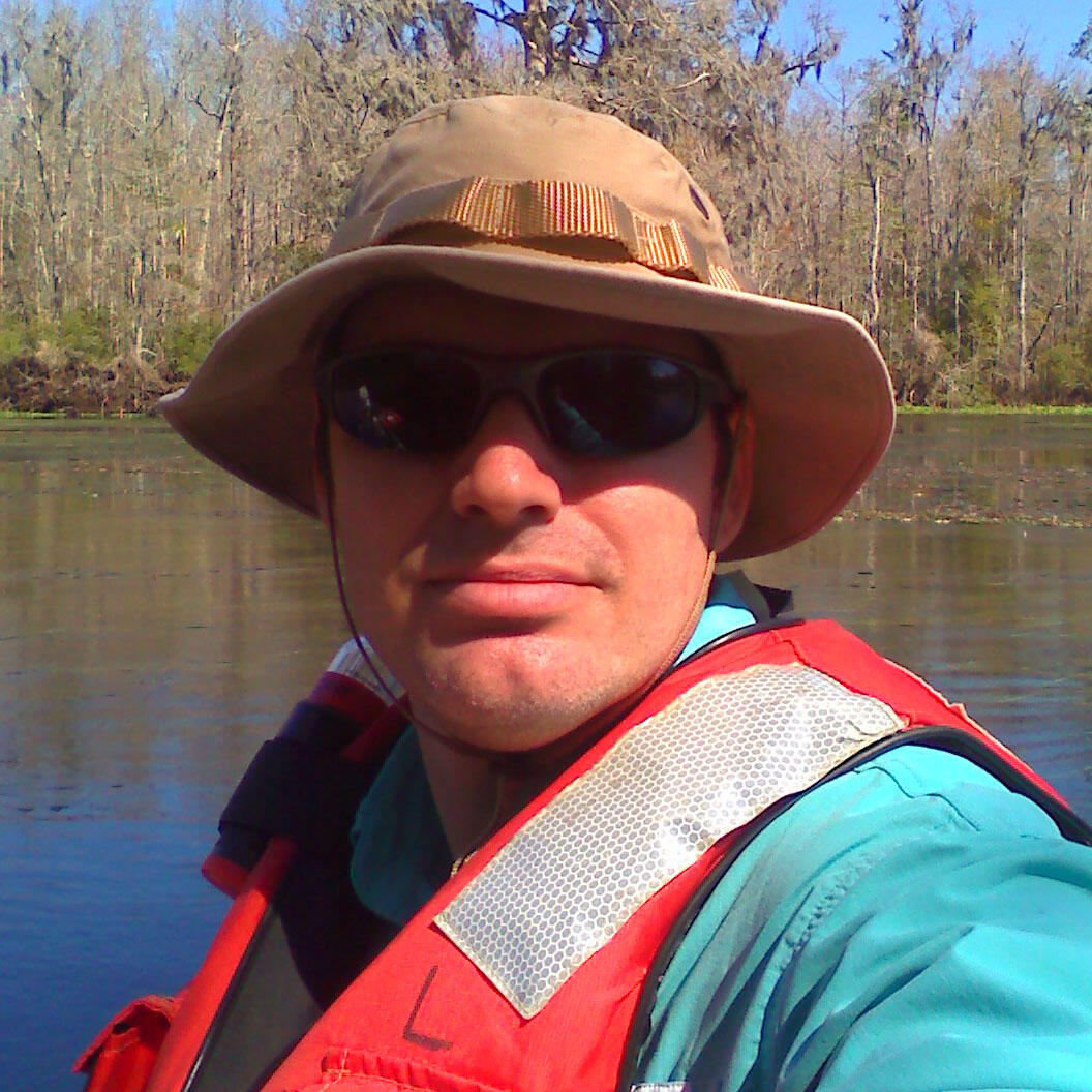 Person with hat, sunglasses, blue shirt, and orange life jacket takes a selfie with water and trees in the background