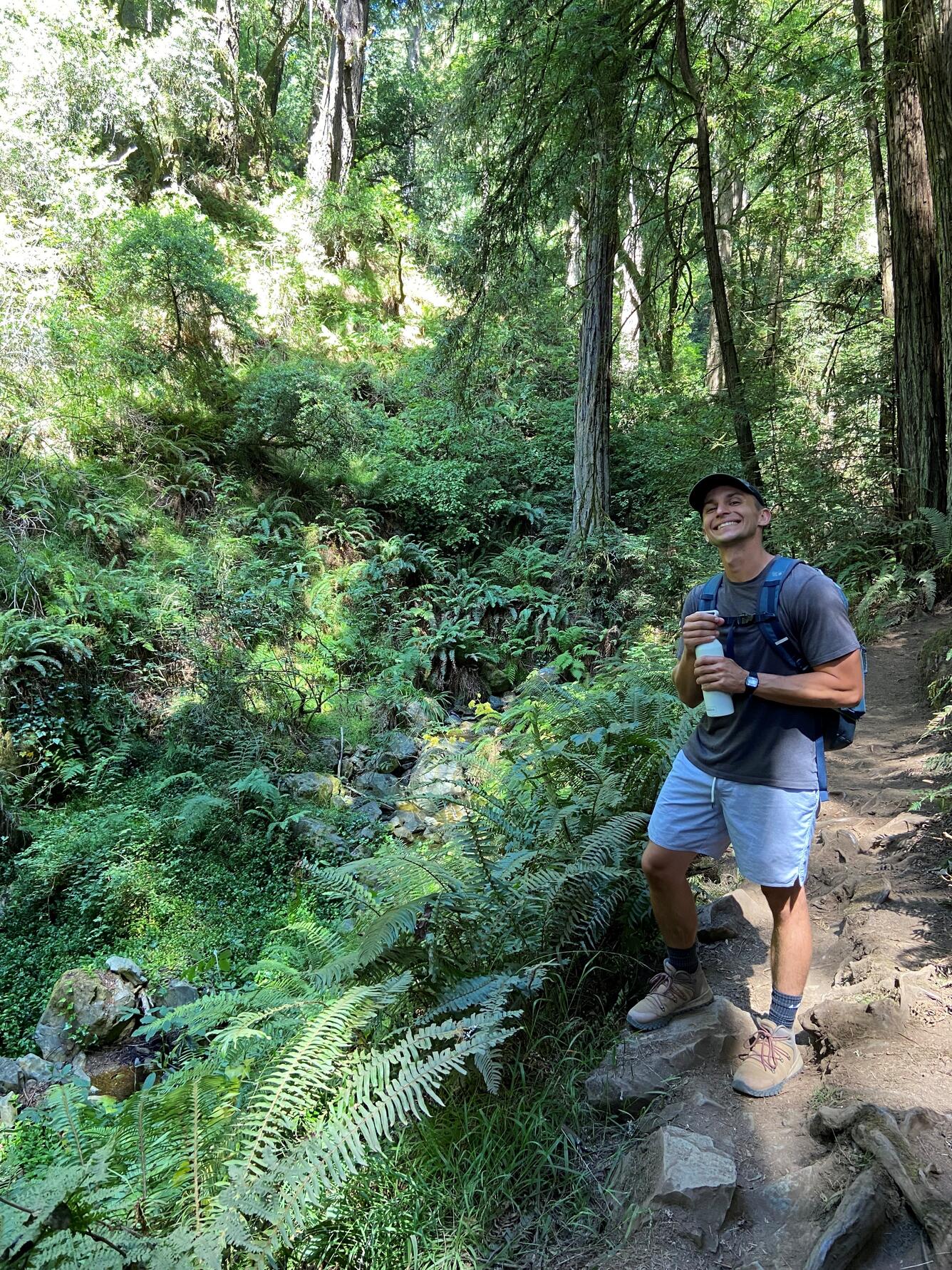 A profile picture of Jackson Valler. He is wearing grey shorts, a grey t-shirt and a black baseball cap as he smiles at the camera while holding a water bottle. He is standing on a hiking trail, surrounded by green, tropical fauna. 