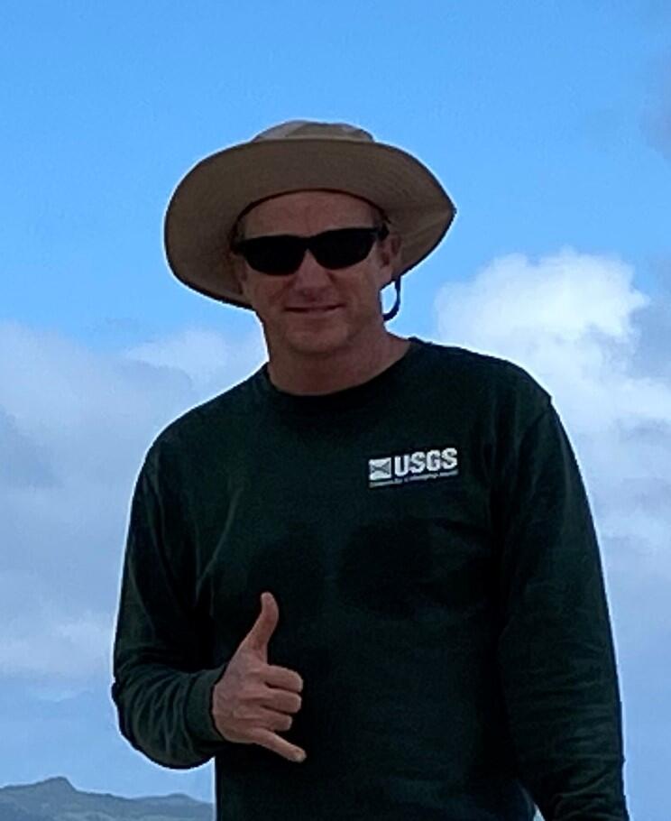 man in long sleeve green USGS shirt and wide brimmed tan hat and sun glasses give the hang loose sign.