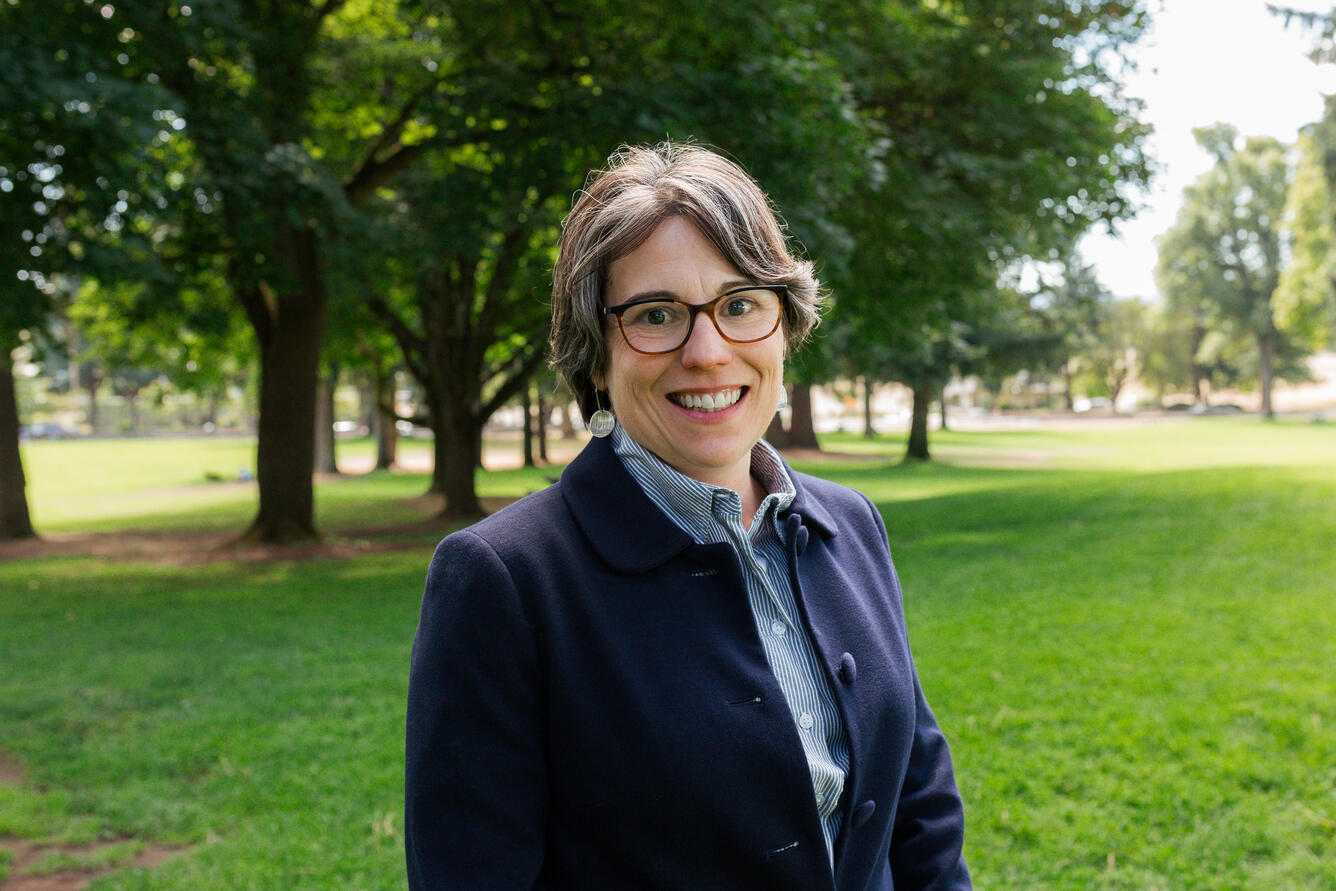 A woman with short brown hair wearing a blue blazer smiles for her picture while in a park.