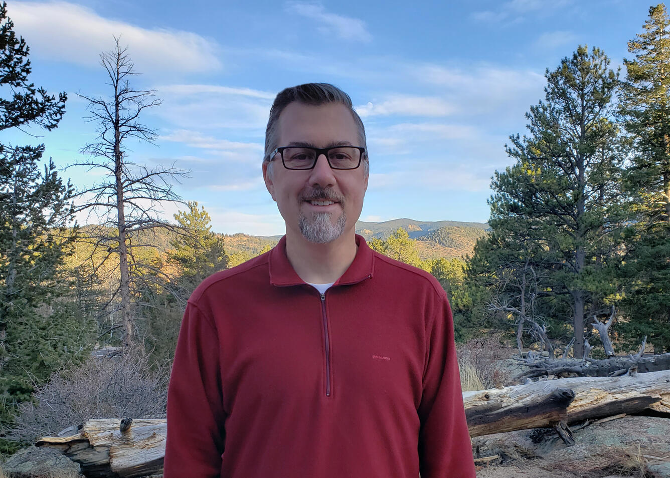 Steve Hanser, smiling and wearing a red quarter-zip sweatshirt, standing in front of trees and mountains in the background. 