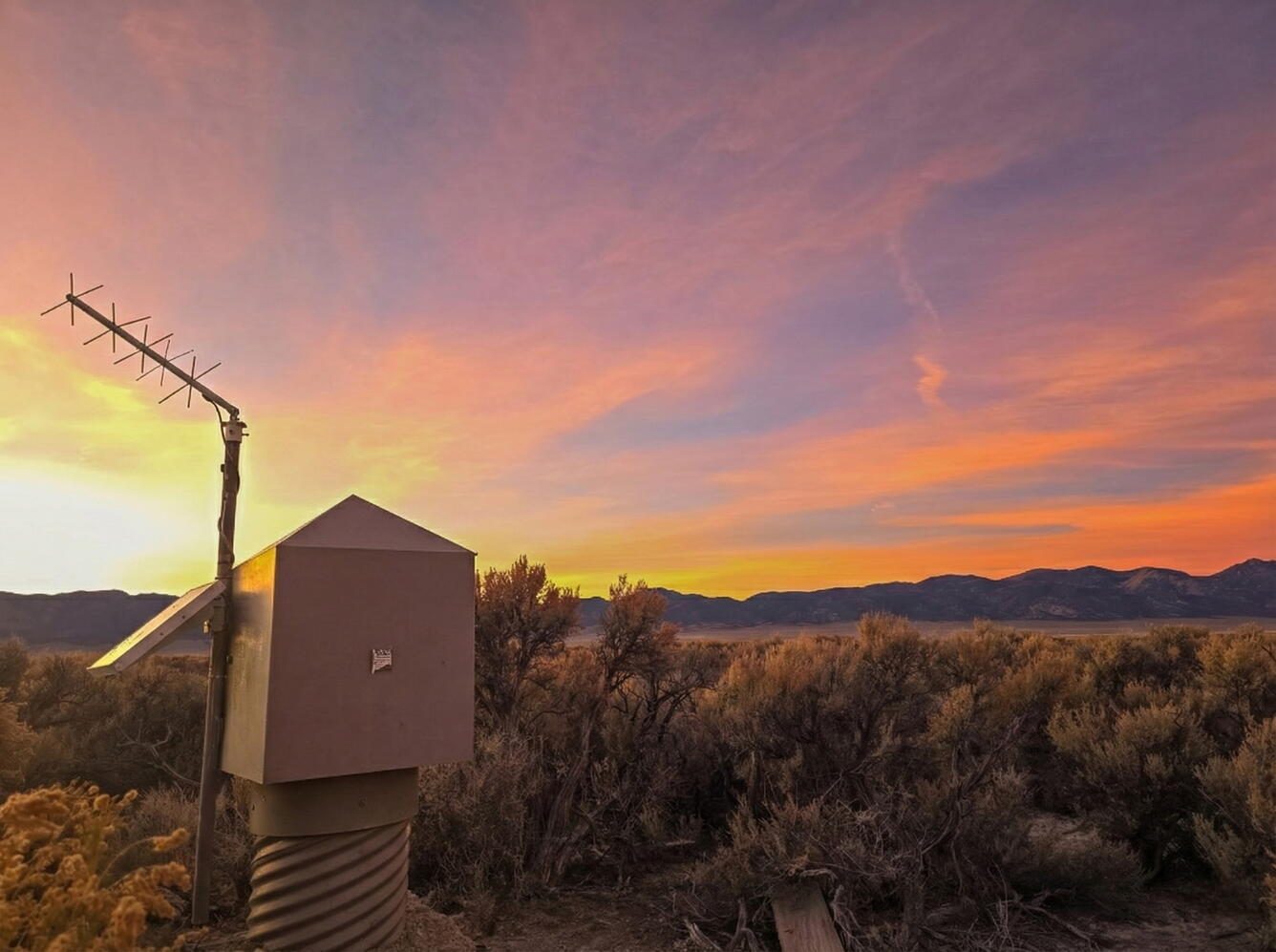 Yellow-orange sunset over desert landscape with groundwater monitoring station and distant mountains.