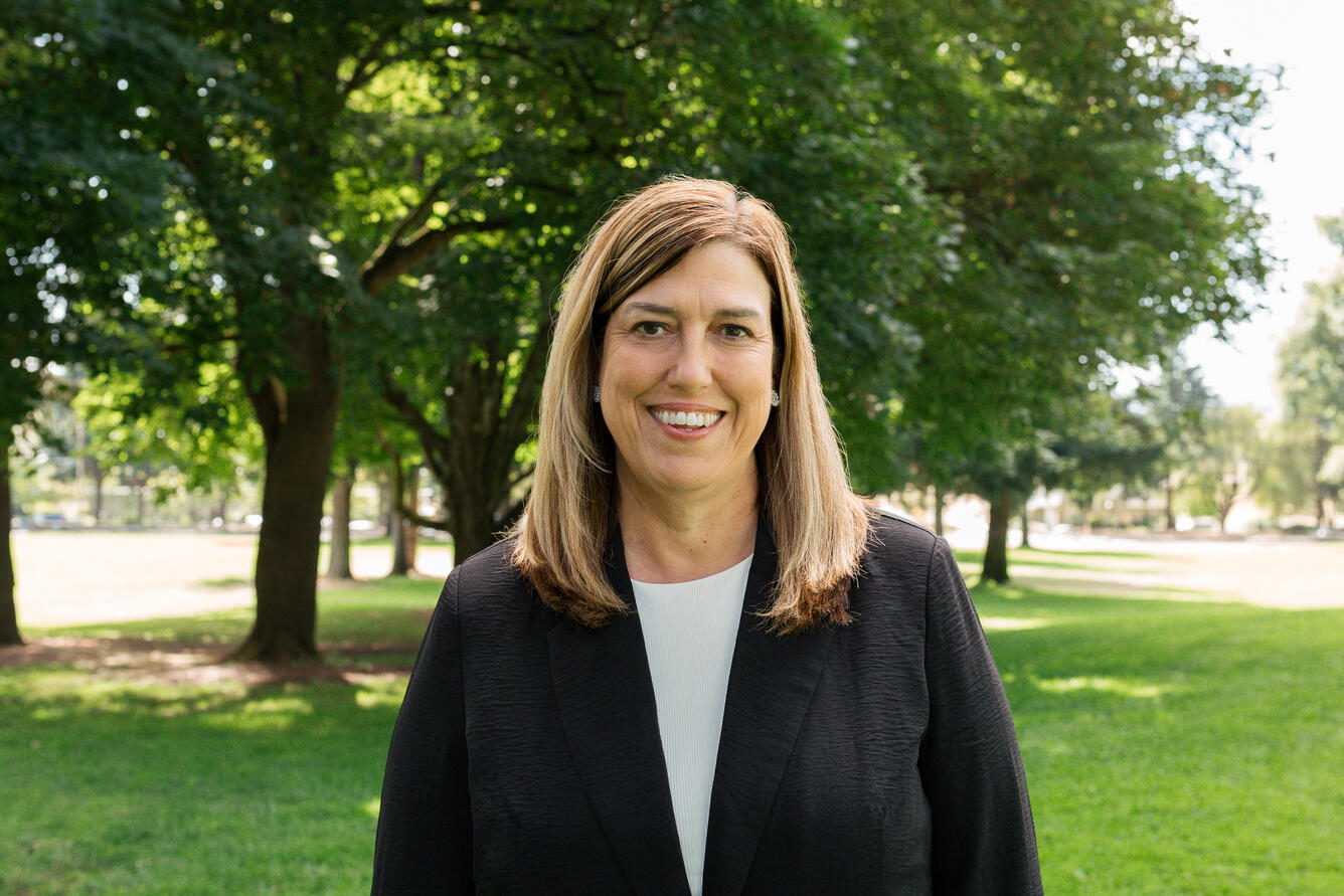 A woman with brown, shoulder length hair wearing a light blue shirt and a black jacket smiles for her picture while in a park.