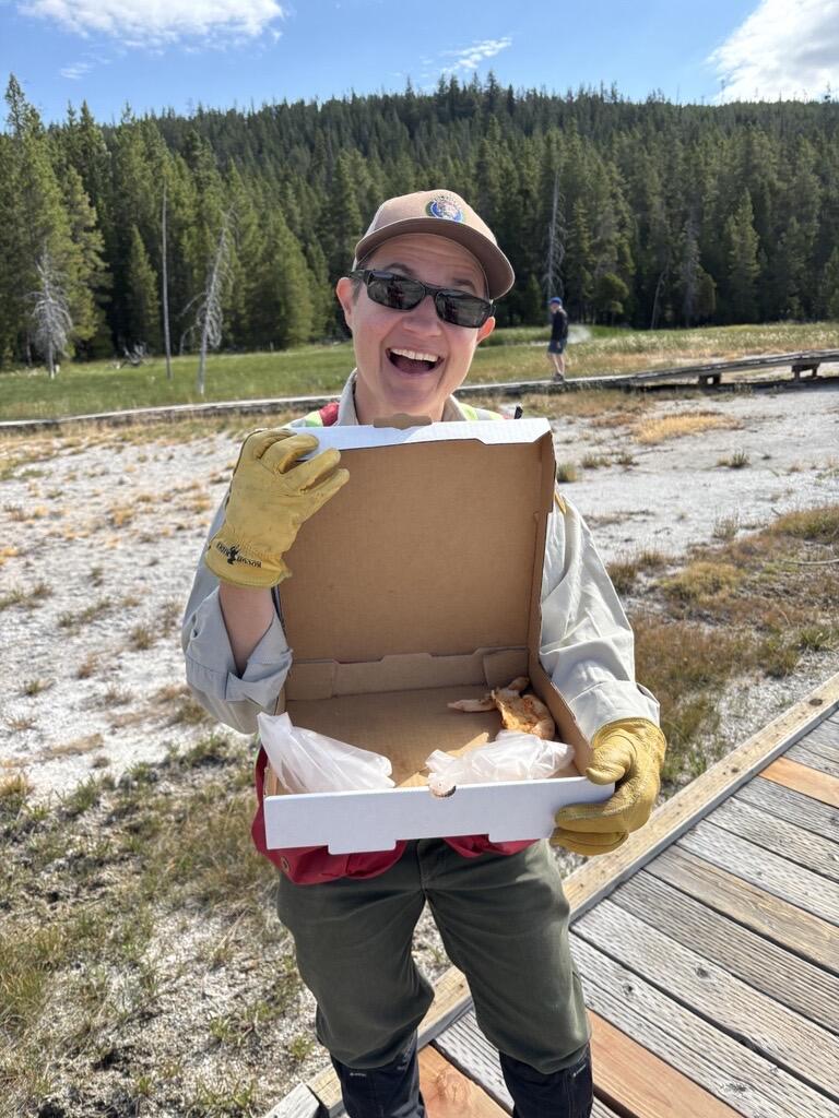 Smiling woman standing on a boardwalk and holding an open pizza box. Scrubby ground and forested hill in background.