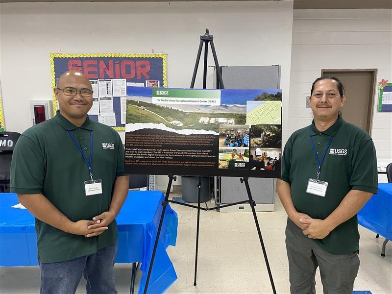 Two people in green shirts stand on either side of a poster inside a community center room.