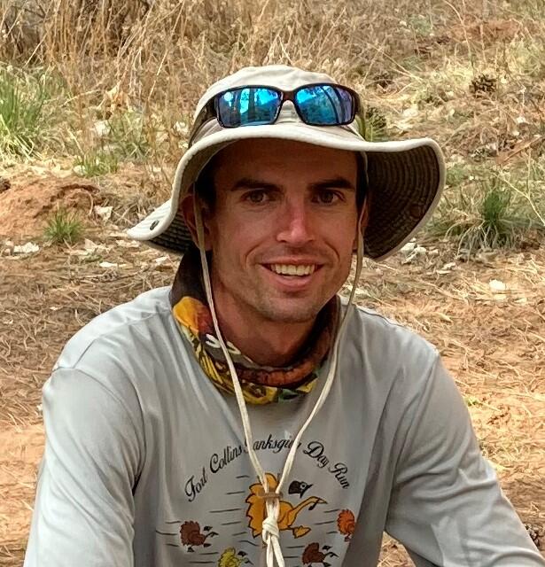 Photo of Tait Rutherford, wearing a brown hat and smiling. There are brown grasses in the background. 