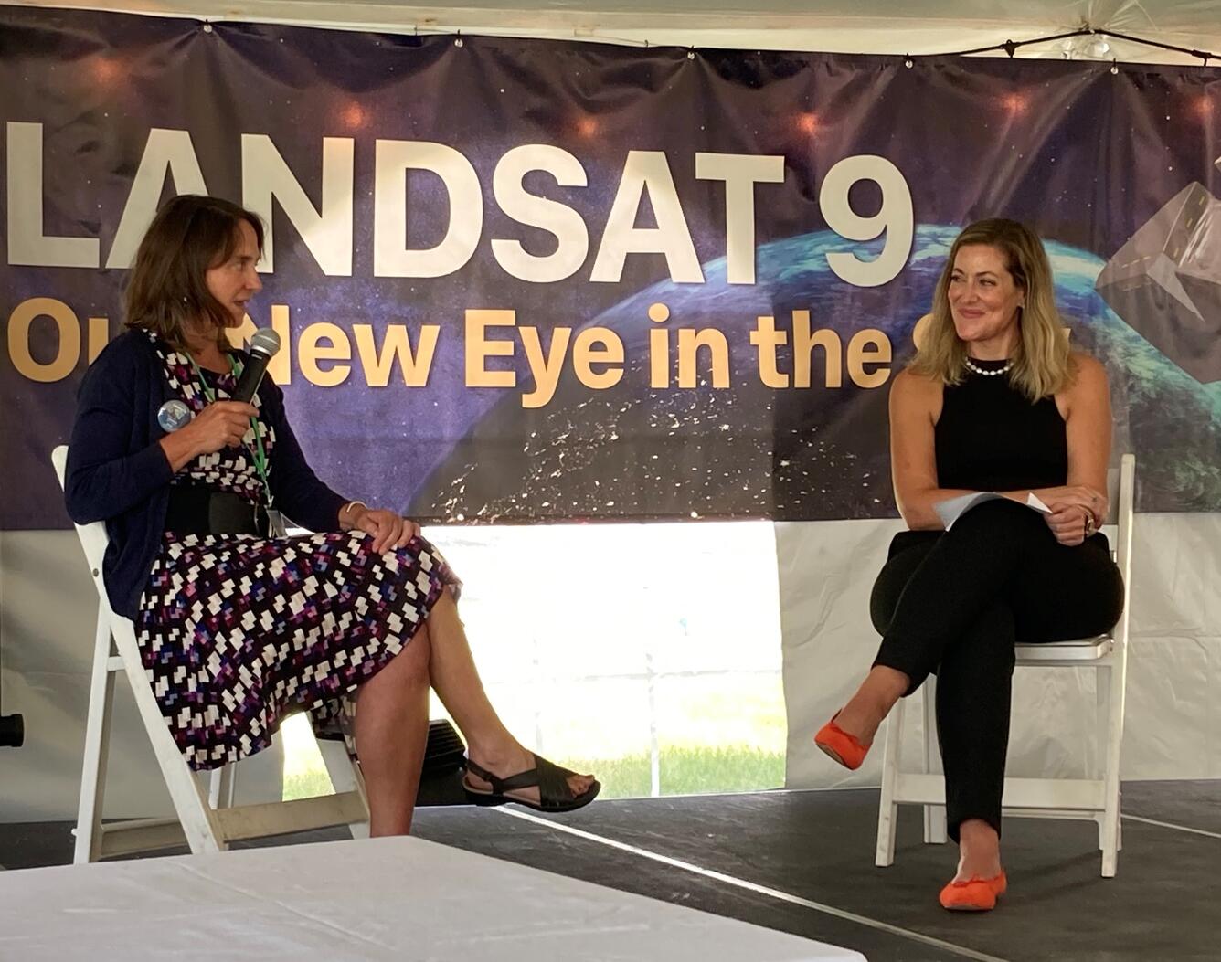 Two women are seated on a stage having a discussion