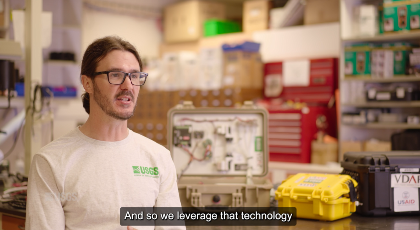 A scientist is sitting in the lab, surrounded by volcano gas monitoring equipment, talking about his work.