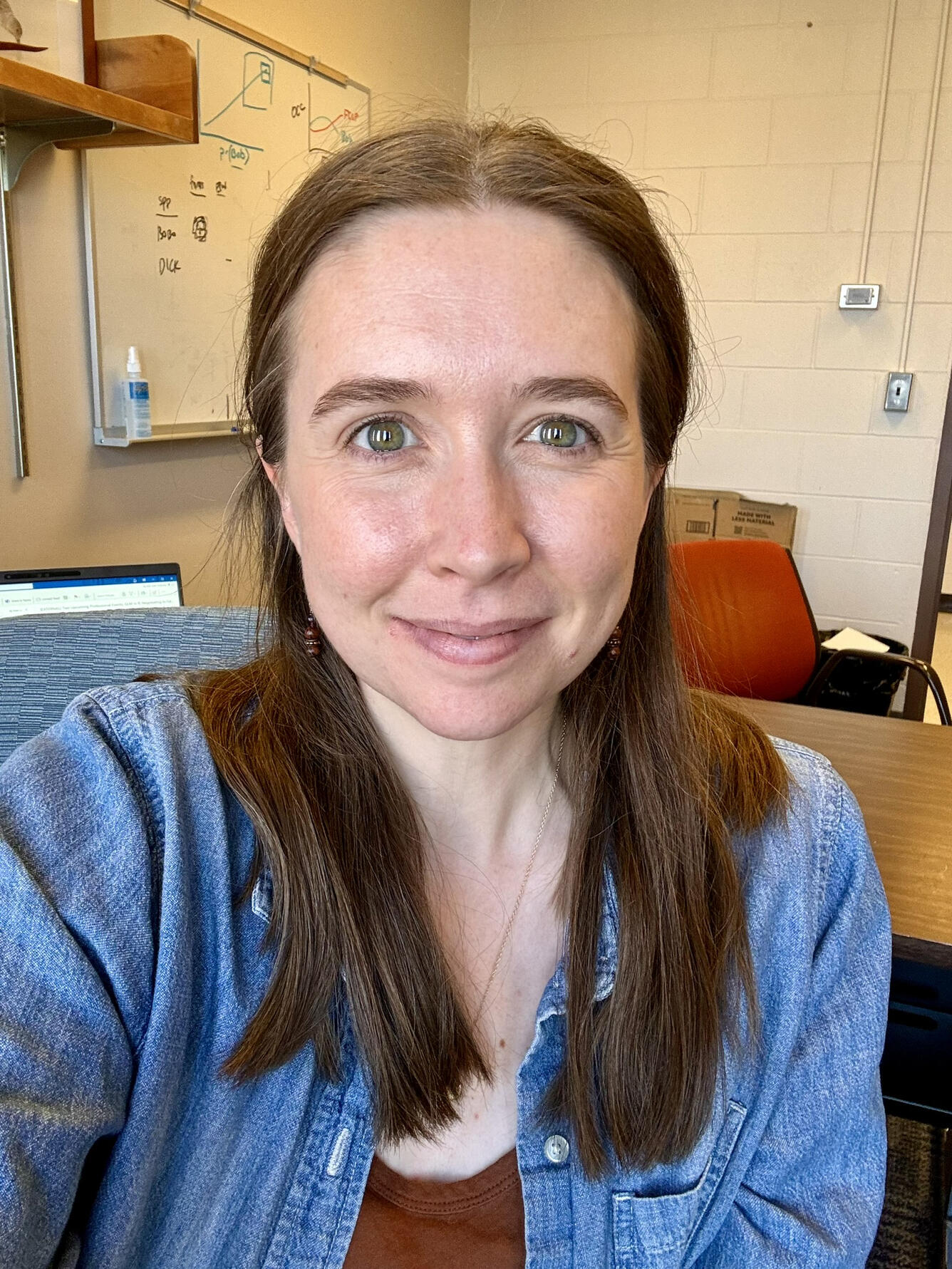 female scientist, smiling, with brown hair, standing in her office near a white board