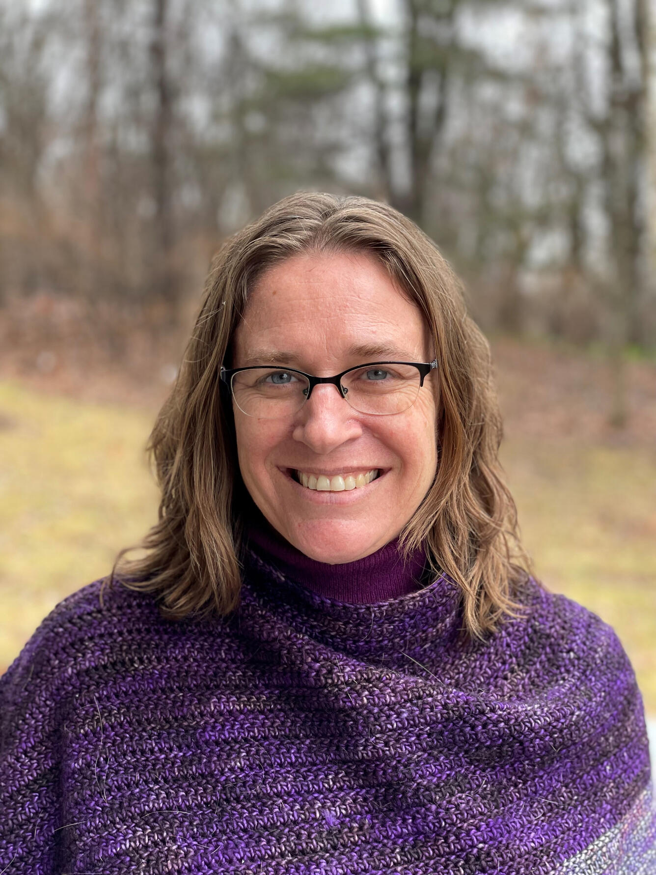 female scientist, smiling, with dirty blonde hair, wearing a purple sweater, standing near the woods