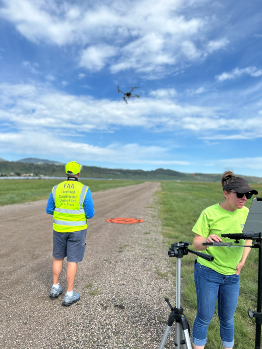 two scientists in bright yellow shirts stand on an airplane landing strip in a field, with a small UAS in the air above them