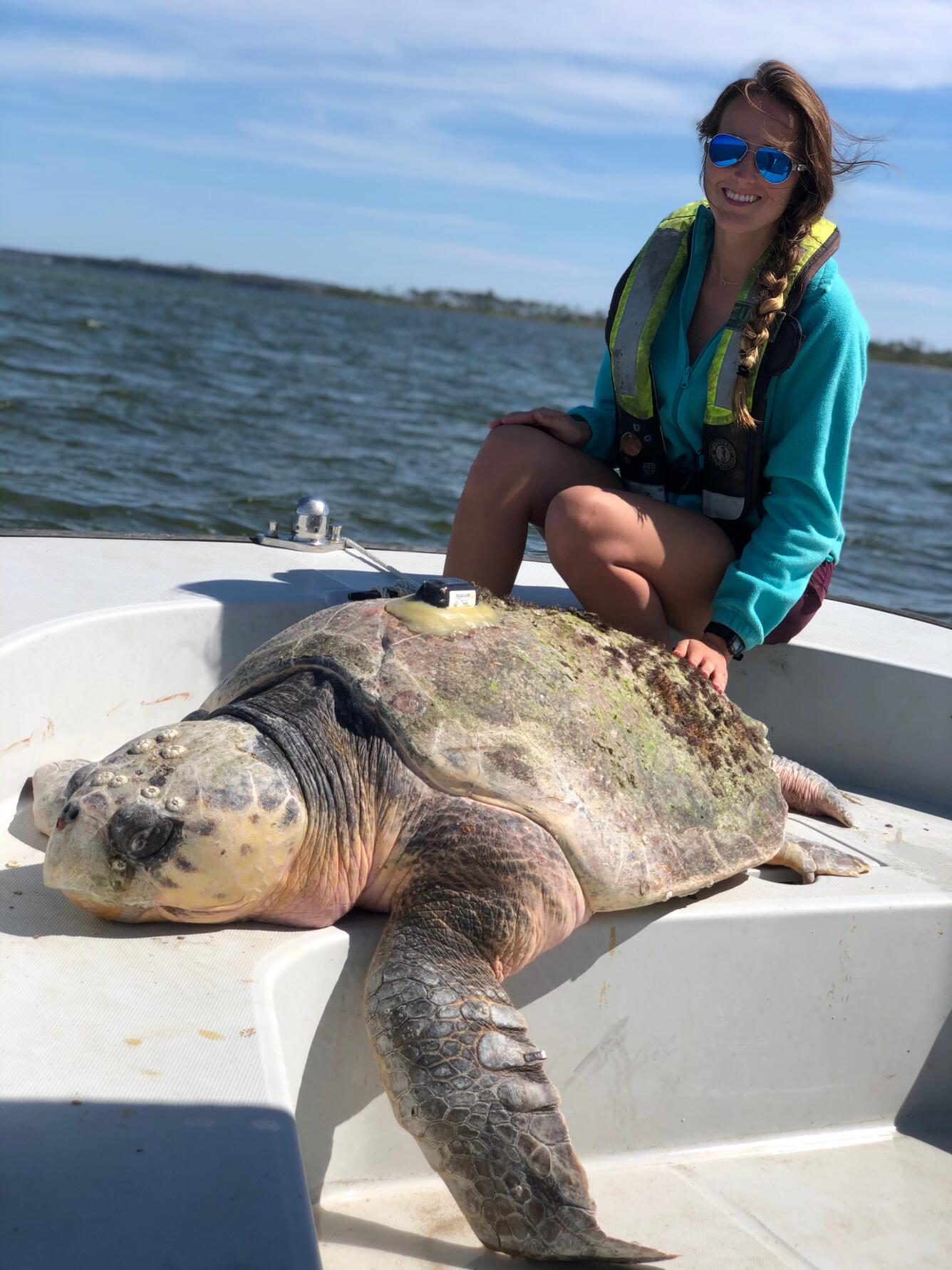a person in a blue shirt and yellow life jacket smiles while sitting at the front of a boat with a sea turtle at their feet