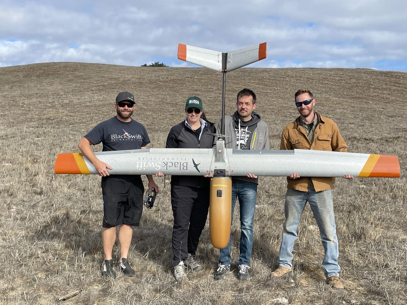 Four scientists stand in an open field holding a large grey and orange drone