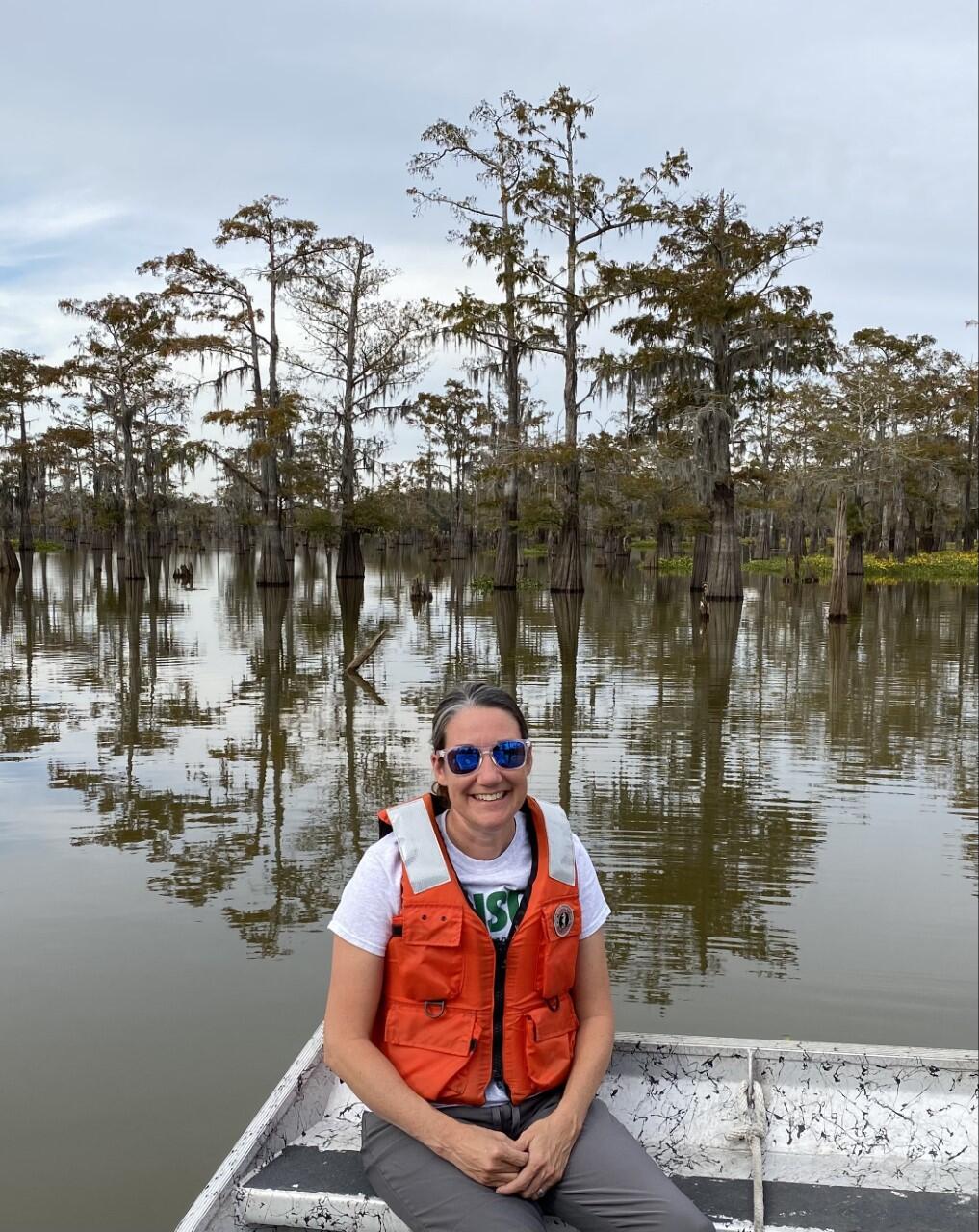 Scientist with orange life jacket by water with bald cypress trees in background