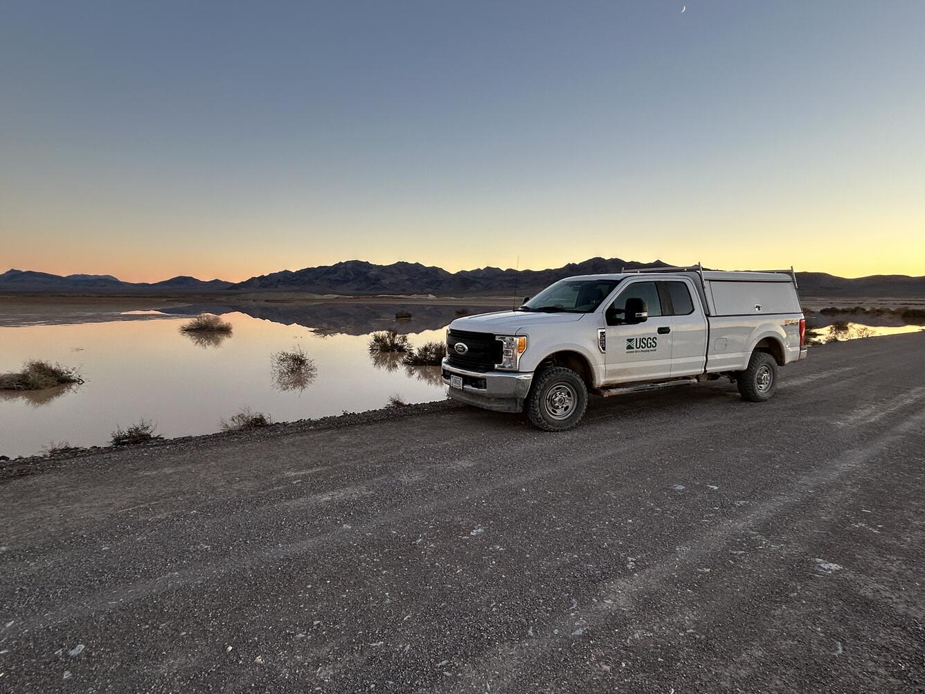 White USGS truck parked on dirt road near shallow-water playa at dusk with silhouetted mountains.