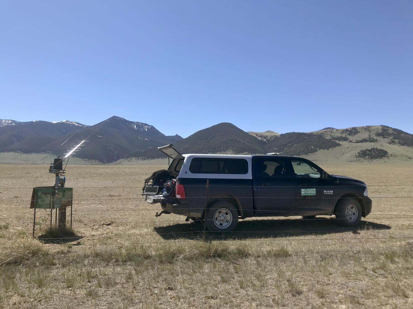 Groundwater well with satellite transmission equipment and a USGS field truck on a wide grassy field with mountains behind.
