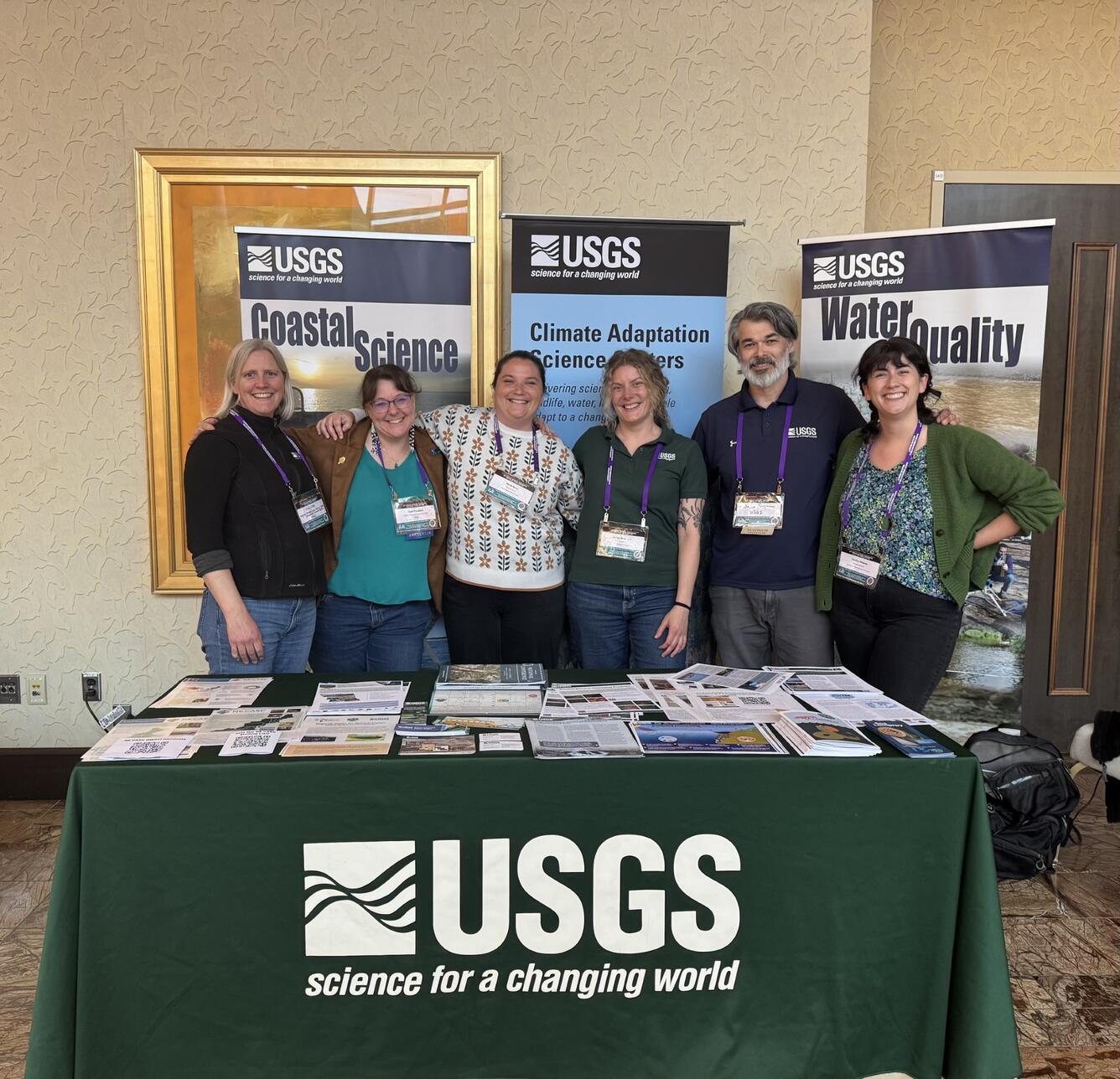 Six people stand at behind a table covered in a tablecloth that has the USGS logo on it.