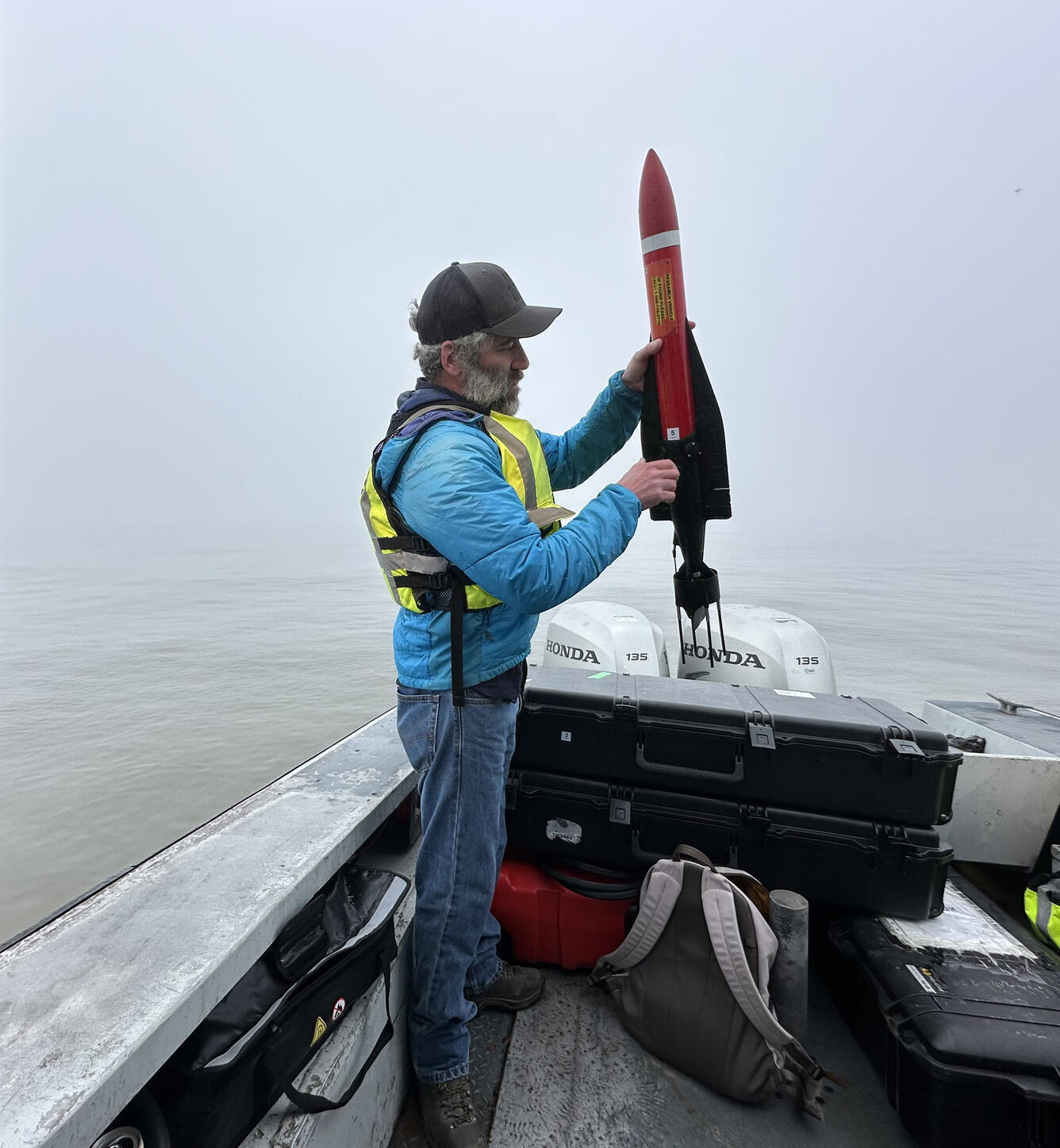 USGS scientist stands in a small boat holding a small, missile-shaped piece of equipment