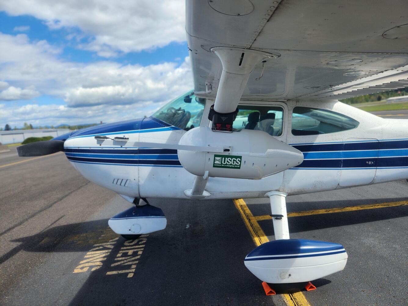 a small white plane with a blue stripe down the middle. USGS sticker on a white enclosure housing Multispectral camera system