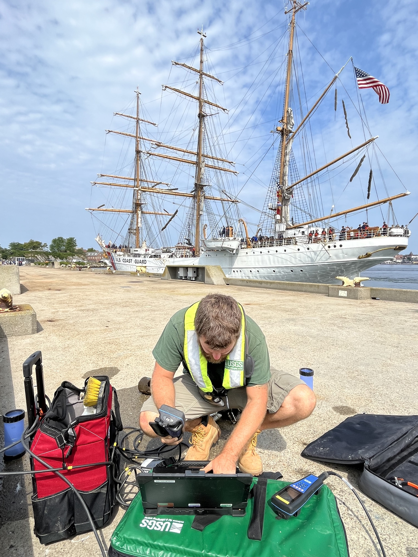 Ryan is crouched down setting up gear on the ground with a large USCG sailboat in the background