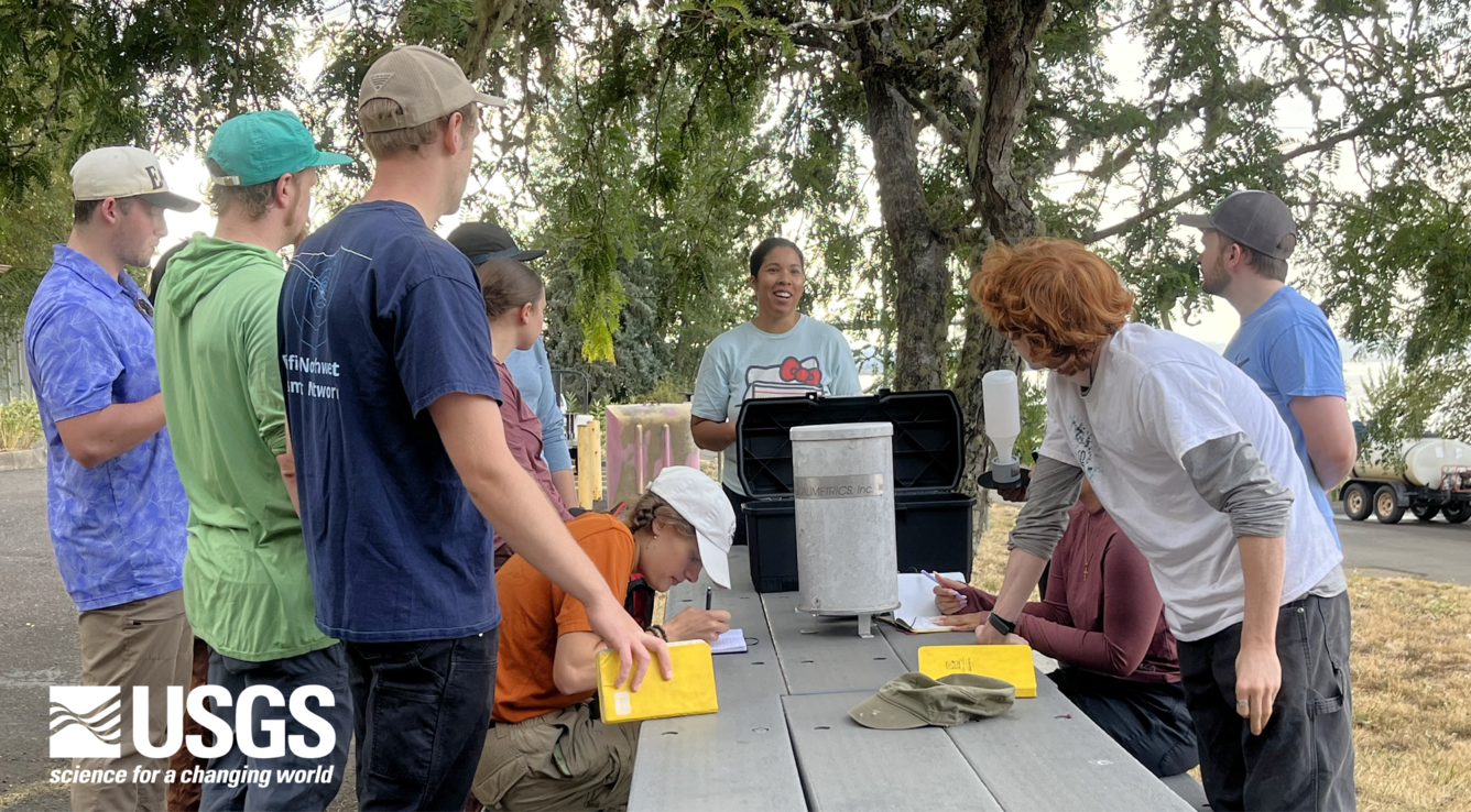 students gather over grey picnic table to look at equipment while note taking in yellow field notebooks