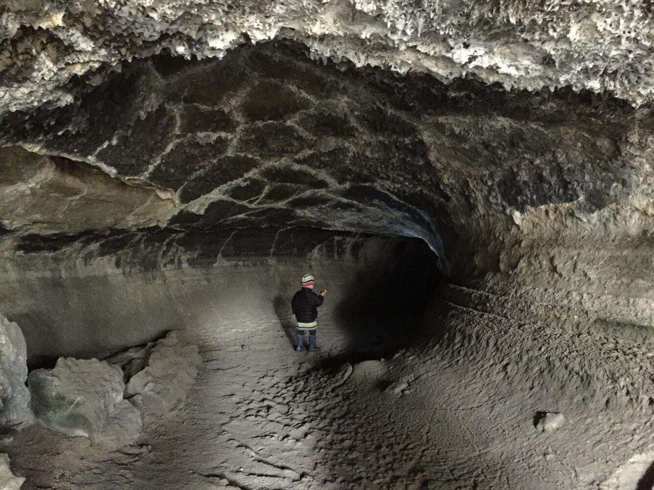 A small child wearing warm winter clothing is viewed from the back, standing in a natural tunnel of dark volcanic rock. The roof of the tunnel is crusted with white minerals and drips of hardened rock, and to the right horizontal raised lines follow the wall of the tunnel to show lava flow levels.