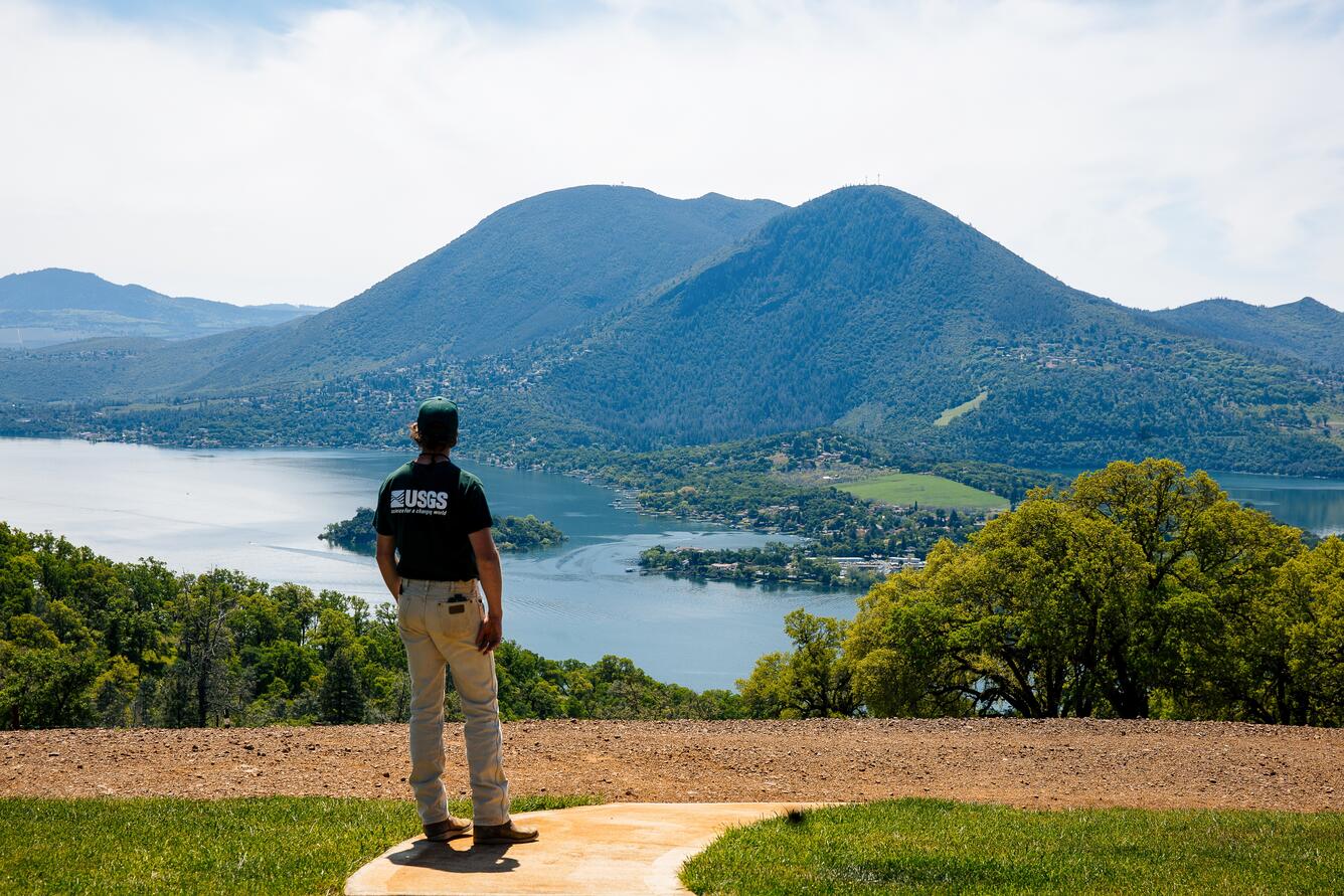 A geologist wearing a green USGS shirt and ballcap is seen from the back, standing at the top of a hill overlooking a lake and a large mountain. The lake is divided by a long peninsula dotted with homes and trees, and the mountain has two prominent peaks that are covered in dark forest.
