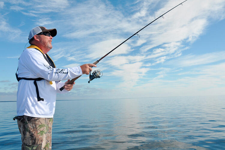 A person in a hat, white shirt, and life jacket holds a fishing pole with water in the background