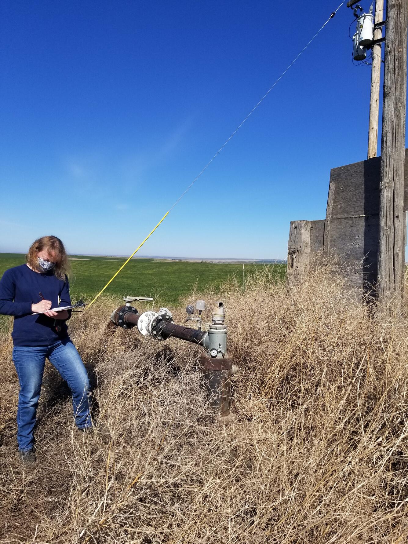 Photo of a person standing in a tall grassy field next to metal piping that is an irrigation well. Person is wearing a mask and writing on a field form on a clipboard. A power line and wooden pole are visible next to them.