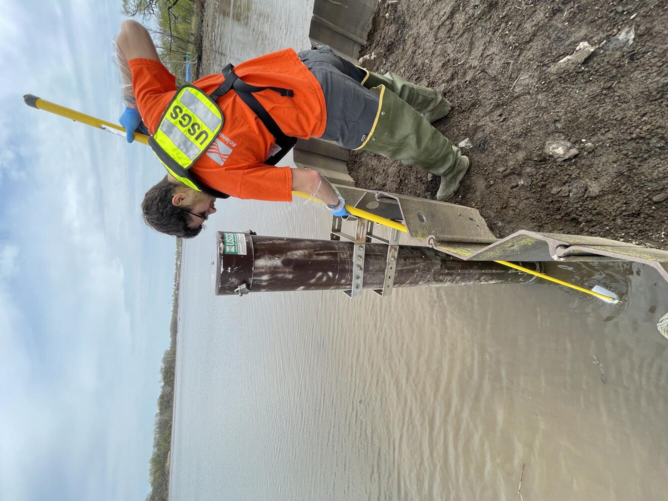 Scientist in Orange and Yellow safety gear uses a long pole to reach into the water and retrieve equipment.