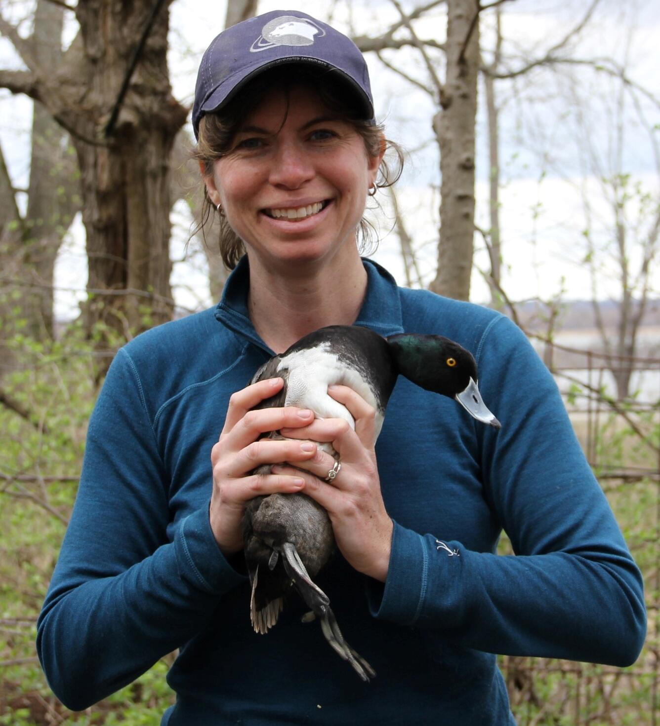 woman with brown hair, blue hat, teal jacket near trees holding Lisa Webb holding a diving duck