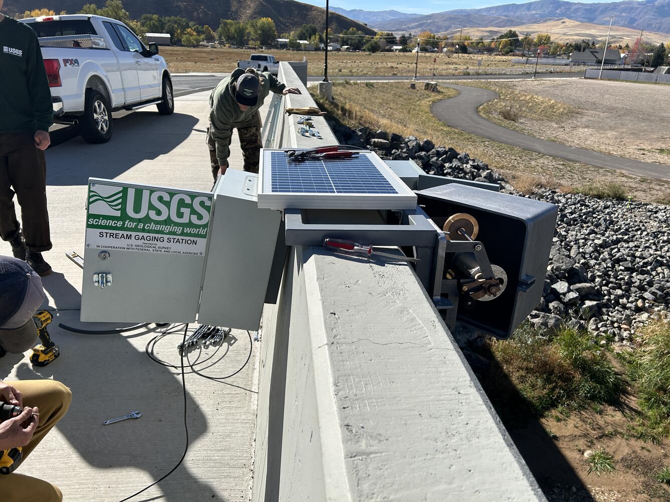 Gage equipment is being installed on side of bridge with partial view of 3 employees.
