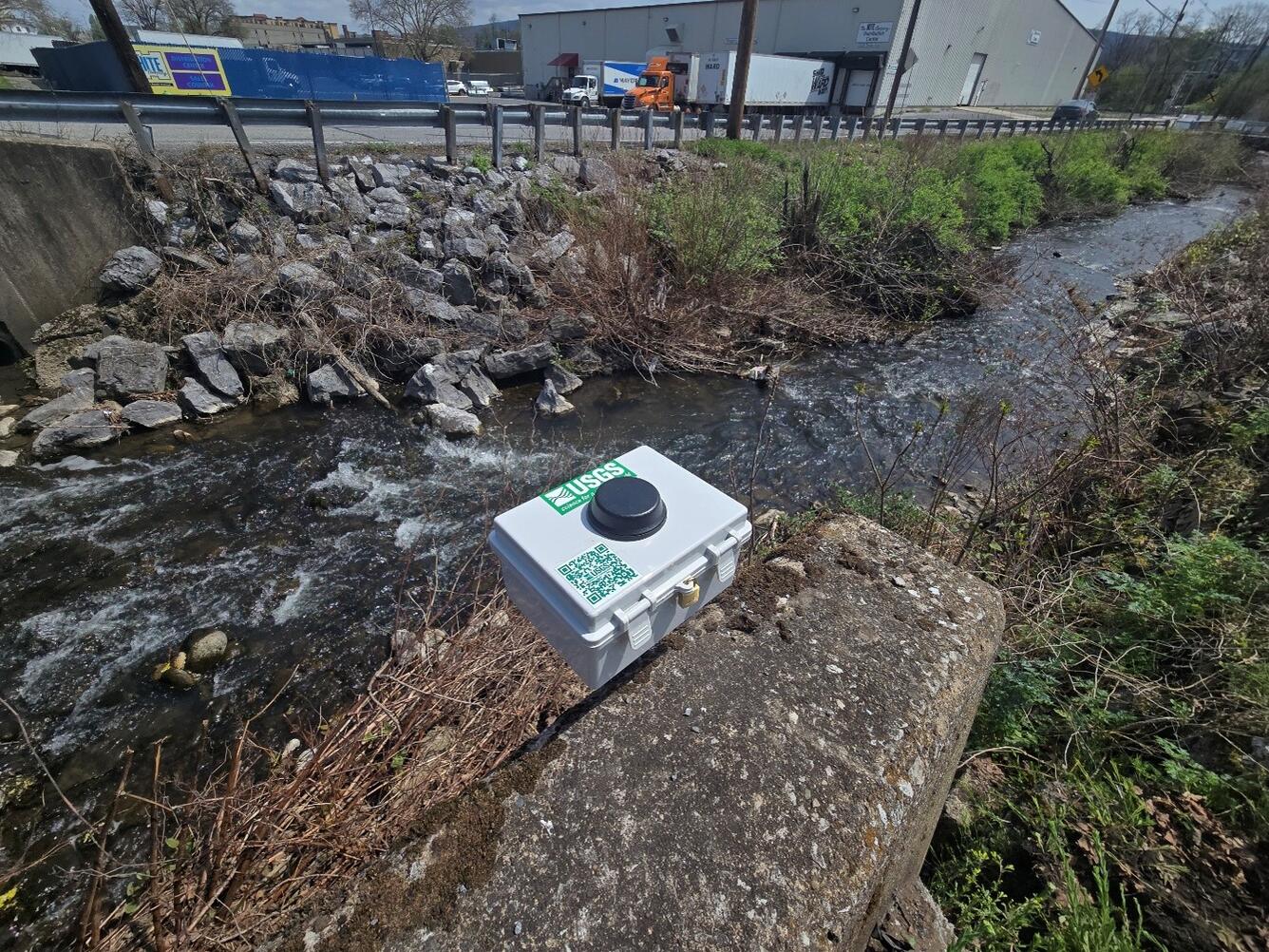 A pressure transducer in a gray box deployed on the downstream wingwall of a bridge